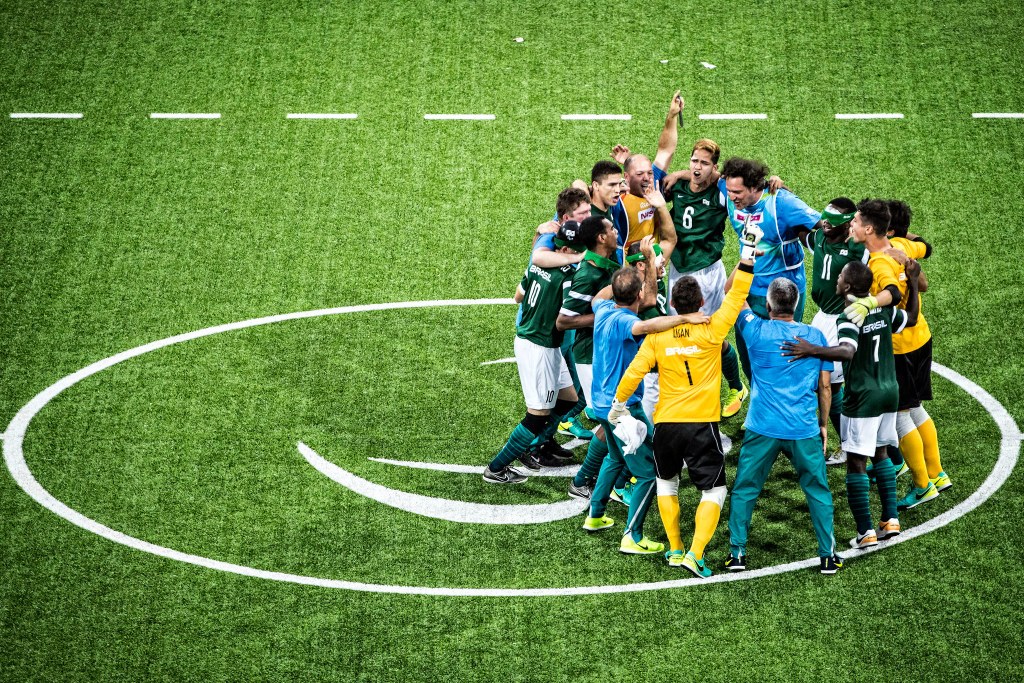 Foto tira de cima. Seleção brasileira de futebol de cinco reunida em no meio do campo verde. Na imagem, aparece 14 homens abraçados comemorando a vitória. Suas expressões são de felicidade e alegria, alguns gritam enquanto pulam com os braços levantados. A farda dos goleiros é blusa de mangas compridas amarela, short preto, meião amarelo e chuteira verde. Os demais jogadores vestem uma blusa que na frente é verde e atrás azul e short branco. Os técnicos usam blusa e calça azul claro.