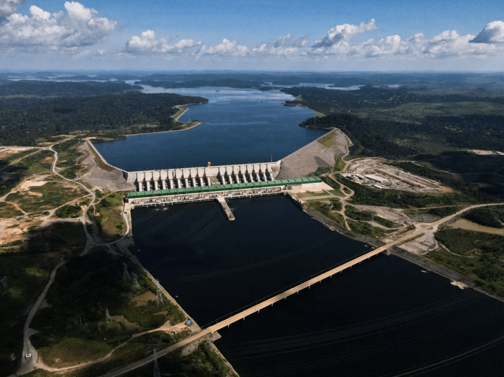 Uma fotografia aérea colorida da Usina Hidrelétrica de Belo Monte, no Pará, Brasil. A imagem mostra a grande barragem de concreto com vertedouros no centro, separando um vasto reservatório de água azul acima de um rio mais estreito abaixo. A paisagem ao redor é composta por colinas verdes cobertas de floresta densa e áreas de terra batida. No canto inferior direito, uma longa ponte atravessa o rio. Uma legenda em texto está presente na parte inferior da imagem.